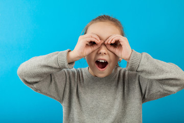 Beautiful girl in surprise looks through binoculars from her hands. Portrait on a blue background.