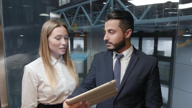 Arabian manager and his Caucasian coworker moving up in lift inside business center and discussing presentation on tablet computer