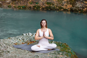 Slim charming blonde woman yoga instructor meditates sitting in the lotus position on a beautiful scenic nature with a lake and stones around. Concept of virgin nature and energy