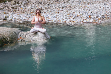 Filling with energy and inspiration from nature. Beautiful young female yoga instructor meditates and relaxes while sitting in the lotus position on a stone near a clean mountain lake
