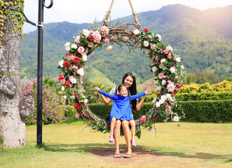 Asian mother and her daughter sitting on beautiful basket swing with the colorful roses flower in the nature garden hanging on pole under tree.
