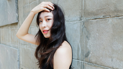 Graceful young woman with long black hair gladly posing with gray wall background. Outdoor close-up photo of cool Chinese girl in trendy summer sexy black dress. 