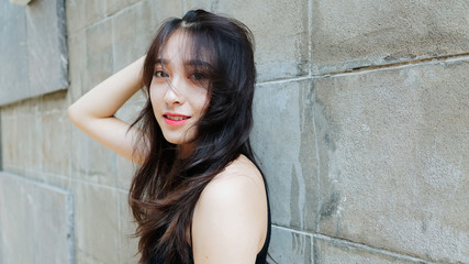 Graceful young woman with long black hair gladly posing with gray wall background. Outdoor close-up photo of cool Chinese girl in trendy summer sexy black dress. 