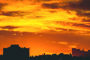 Cityscape with vivid fiery dawn. Amazing warm dramatic cloudy sky above dark silhouettes of city buildings. Orange sunlight. Atmospheric background of sunrise in overcast weather. Copy space.