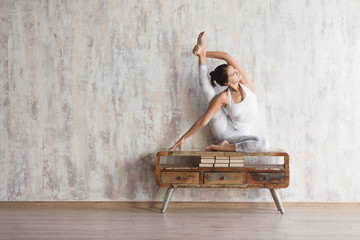 Beautiful young woman yoga instructor doing asana in a white suit on a wooden vintage console in a spacious bright gymnasium. Space for text