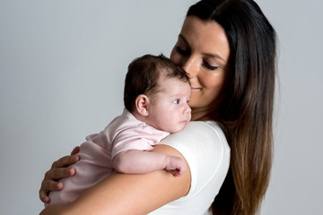 A beautiful young mother standing and holding her baby girl in her arms in front of a grey background in a studio.