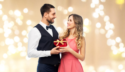 valentines day and people concept - happy couple with gist box in shape of heart over festive lights background