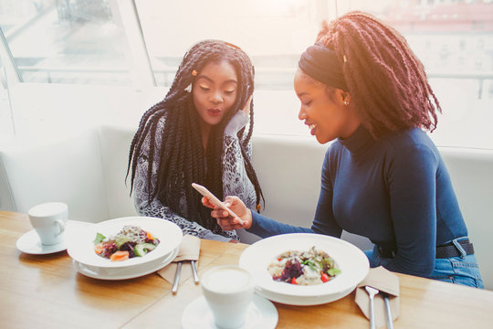 Two african american women sit in cafe at table. They look at phone. Food in balls are on table.