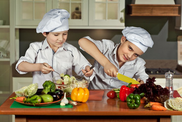 The boys in the cooks costumes cut the salad in the kitchen. Children prepare healthy food. Friendly cooperation of cooks.