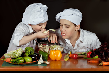 The boys in the cooks costumes cut the salad in the kitchen. Children prepare healthy food. Friendly cooperation of cooks. Isolated on black background.