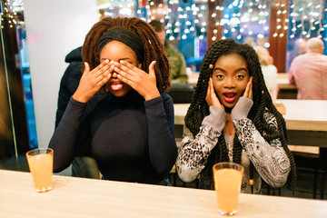 Cheerful nice young african women sit at table and have fun. First cover eyes with hands. Second keep ears closed. Glasses with orange juice stand at table.