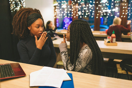 Two young african women gossip at table. On of them looks wondered. Another model keep finger at mouth. They have laptop and study materials on table.