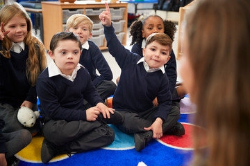View over female teacher's shoulder of primary school kids sitting on the floor in the classroom...