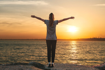 Fototapeta premium A girl in solitude stands on the shores of the Bosphorus in Istanbul in Turkey and raises her hands up showing how she is free and how she likes. There is a beautiful sunset ahead.