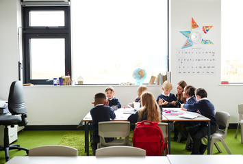 A group of primary school kids sitting at table in a classroom, teacher kneeling to help them...