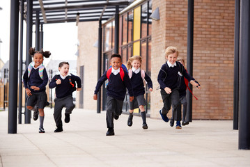 Primary school kids, wearing school uniforms and backpacks, running on a walkway outside their...