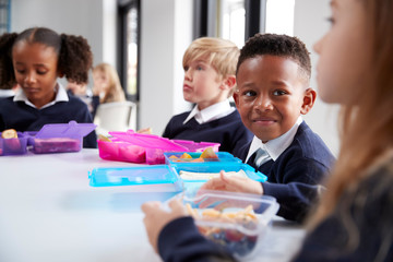 Smiling primary school kids sitting at a table eating their packed lunches together, selective focus