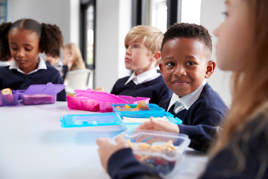 Smiling Primary School Kids Sitting At A Table Eating Their Packed Lunches Together, Selective Focus