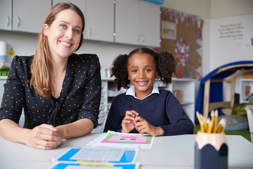 Young female primary school teacher and schoolgirl sitting at a table, working one on one in a...