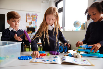 Fototapeta premium Three primary school children working together, following an instruction book and using construction blocks in a classroom, front view, close up