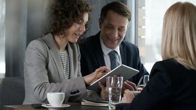 Experienced middle aged businessman wearing formal suit and glasses sitting at restaurant table with young female colleague and explaining something to partners during lunch