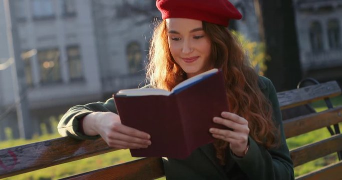 Portrait of the Caucasian beautiful youg girl with red hair and in a cap resting on a bench outdoors with a book, then looking at the side and dreaming.
