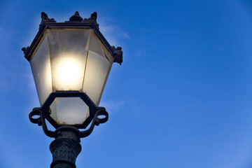 Victorian Style Lamp Post with Lighted Bulb and Broken Glass Panel. Upward Shot of Outdoor Street Lantern made of Black Wrought Iron Cast under a Blue Sky