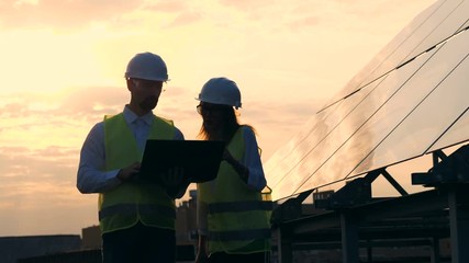 Two workers look at a laptop, walking near solar panels, close up.