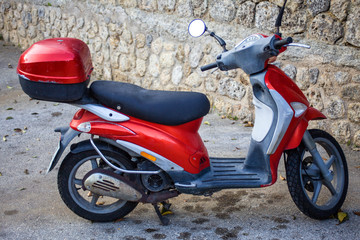 Fototapeta na wymiar One Red Motorbike with Flat Floor Board Step Parking on a Dirty Lot. Sideview Shot of Motorcycle with Helmet Storage Case Facing an Old Stone Paved Wall