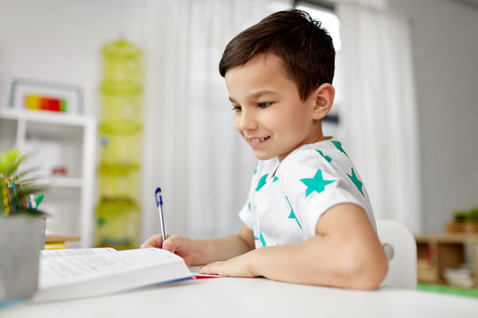 education, childhood and people concept - happy student boy with book writing to notebook at home