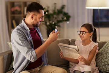 family, technology and people concept - happy father photographing daughter reading newspaper by smartphone at home