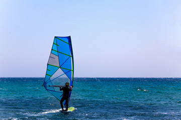 Rider in Wetsuit and Helmet Balancing on a Surfboard with Sail Mast and Bar. Clear Sky and Blue Wave. Surfer Trying out the Outdoor Water Sport Adventure