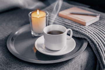 Cup of  hot coffee with a candle on a gray tray, next to a knitted gray scarf and a white sweater. Concept cozy winter home relax