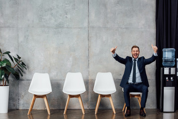 excited businessman in suit sitting and showing thumbs up in waiting hall