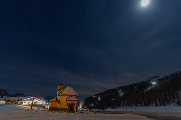 Night view of a small church at the San Pellegrino Pass in winter, with the snow-covered ski slopes in the background, Dolomites, Italy