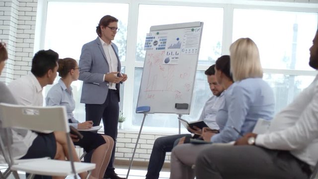 Male business coach writing on flipchart while giving presentation to colleagues sitting in seminar room and writing down in notepads during corporate training