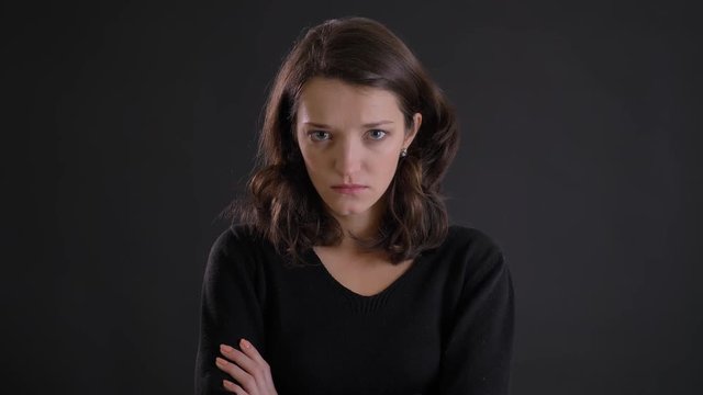 Portrait of young beautiful caucasian long-haired girl with crossed arms and watching angrily into camera on black background.
