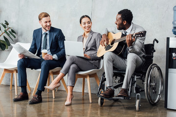 Obraz premium multicultural businesspeople sitting and listening while disabled african american man playing guitar in waiting hall