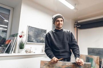 Indoor portrait of attractive young artist, standing in the studio and posing on the camera. Young artist with a picture in his hands stands and smiles.