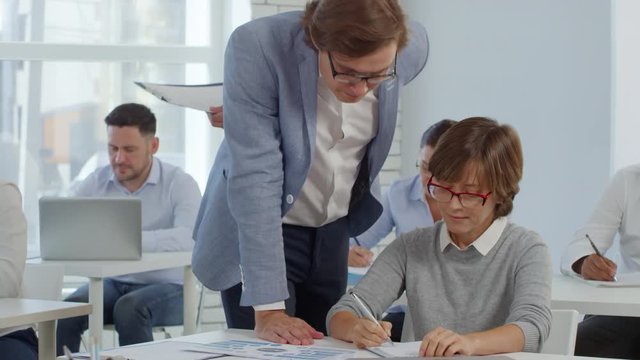 Tracking shot of office workers sitting at desks and writing down while taking test during corporate training program; male coach helping middle aged businesswoman