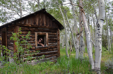 Woman urban explorer smiles through a broken window in an old log cabin in Miners Delight Wyoming