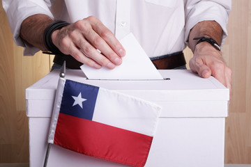 A Chilean citizen inserting a ballot into a ballot box. Chile flag in front of it