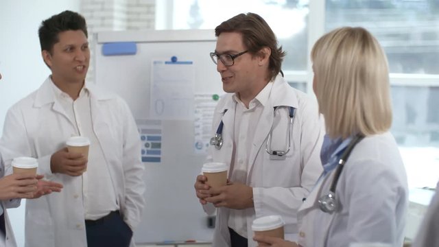 Cheerful male physician talking with multiethnic colleagues in lab coats while having coffee break in clinic