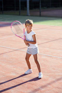 Little female tennis player in white uniform concentrating and focusing on the training before game. Sunlight background.