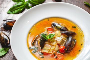 Soup with mussels and vegetables in white plate on wooden table. Close up