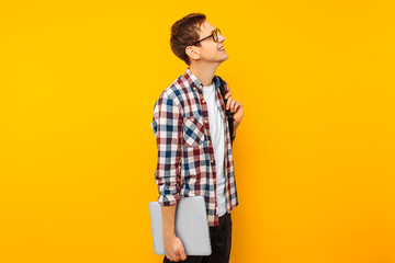 Happy guy in glasses, a student in a plaid shirt, goes with a closed laptop in his hands, on a...