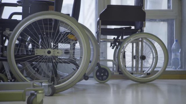 Slow motion of bunch of new wheelchairs with white wheels put in raws next to big windows at daylight in hospital