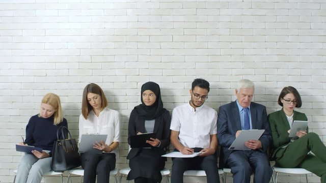 Medium shot of group of diverse job candidates sitting in row in office reception area and chatting while taking skills assessment tests before interview