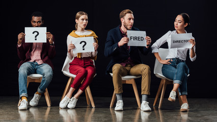 multiethnic people sitting on chairs and holding cards with 'director', 'fired' words and question marks isolated on black