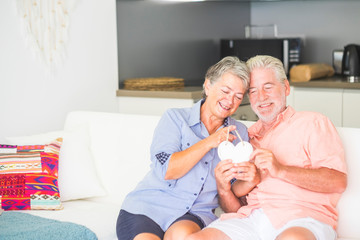 Bright image couple of man and woman caucasian people happy at home sitting on the couch with kitchen in background playing with white hearth together with love - valentine's day concept for elderly